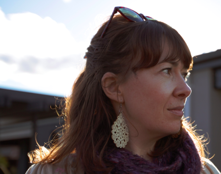 Phoebe is looking into the distance. The sun shines behind her. Phoebe wears earrings, a scarf, and sunglasses on top of her head. 