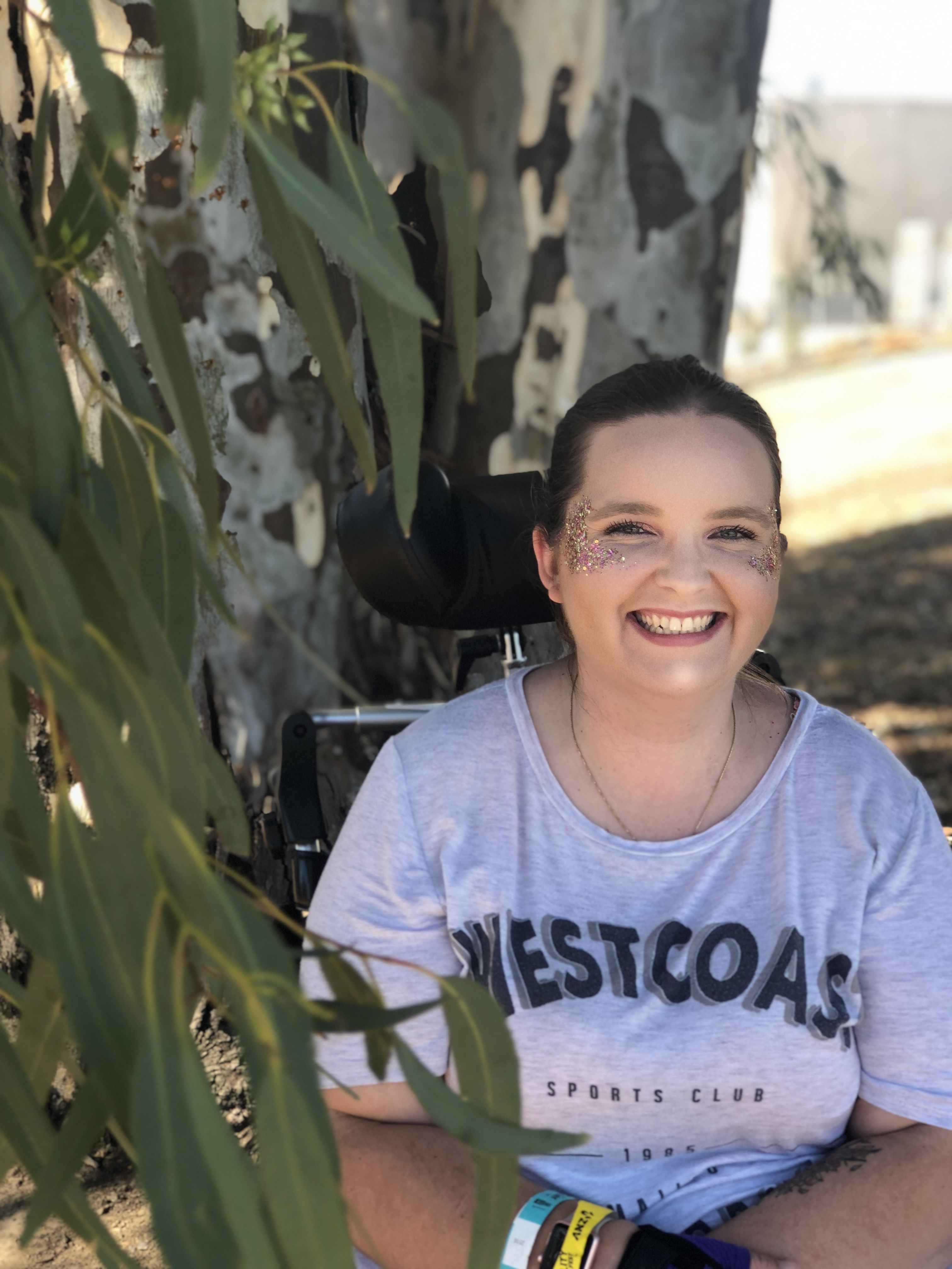 Jess has glitter on her cheekbones and is smiling widely. Gum leaves are dangling near Jess' face. Jess is a wheelchair user.