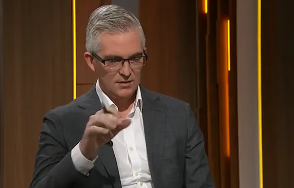 David Speers is talking.  He wears glasses and his right hand is raised 