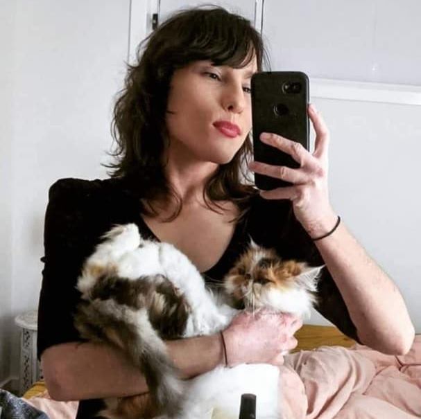 Elena is taking a photo in the mirror. She's holding a smartphone, and is looking at the phone's screen. Elena is also cradling a cat. 
