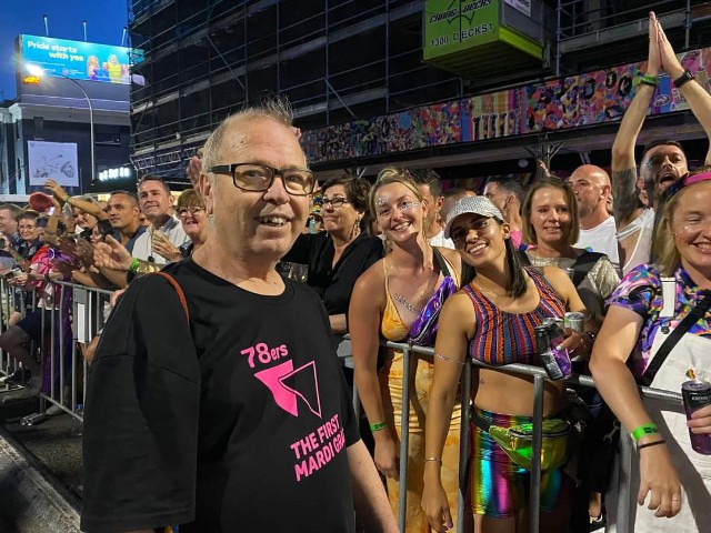 Image shows: Dr David Abello with a crowd of people at Mardi Gras/World Pride parade in 2023. Everyone is smiling and looking at the camera. David is wearing a black t-shirt with pink writing on it reading: '78ers - The First Mardi Gras'. 