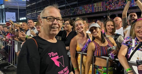 Image shows: Dr David Abello with a crowd of people at Mardi Gras/World Pride parade in 2023. Everyone is smiling and looking at the camera. David is wearing a black t-shirt with pink writing on it reading: '78ers - The First Mardi Gras'.