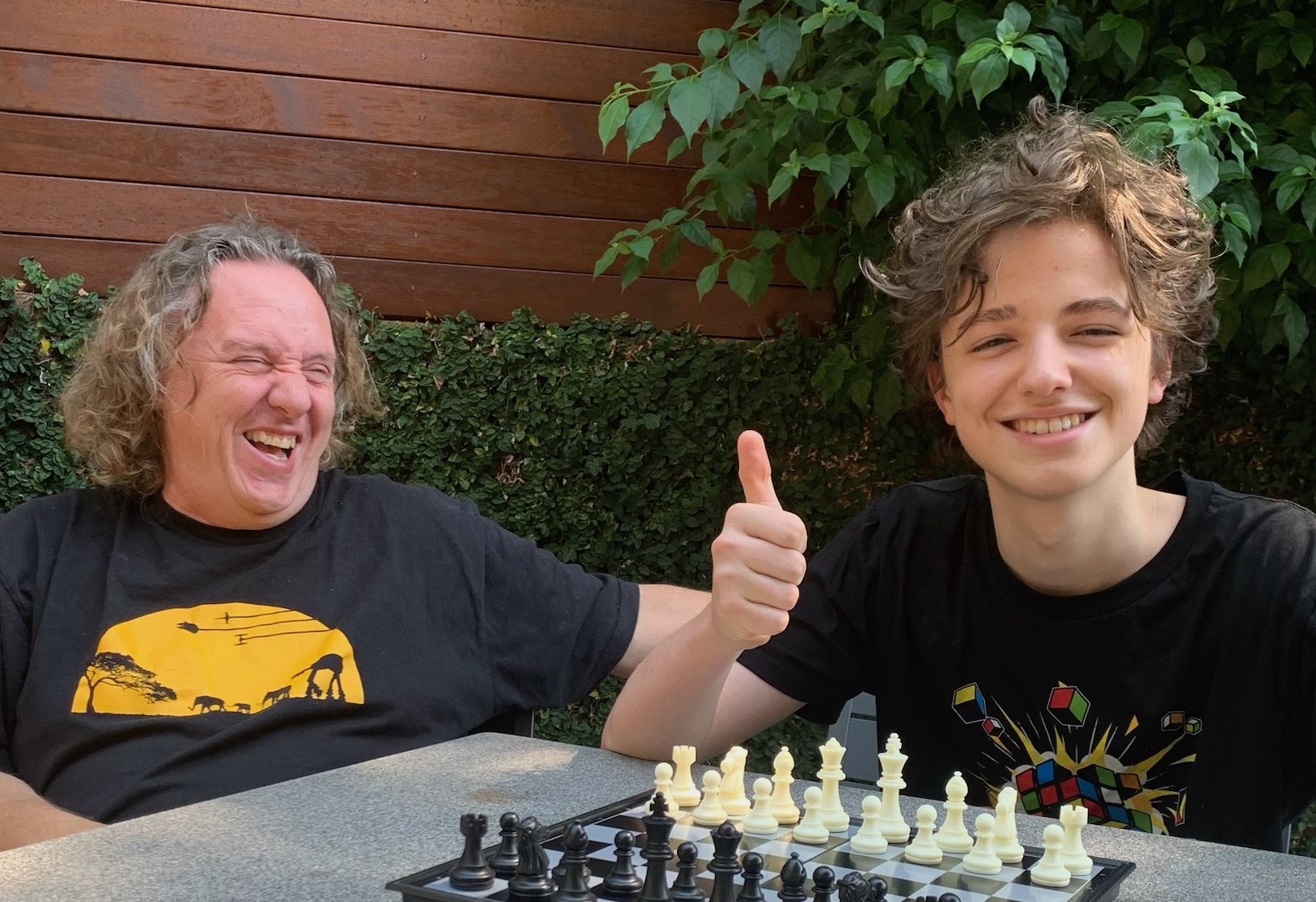 Peter and Rafferty are outside playing chess and smiling. Peter has long, light brown hair and is wearing a black t-shirt with a yellow sun image. Rafferty has short brown hair and is wearing a black t-shirt with a rubik's cube image.