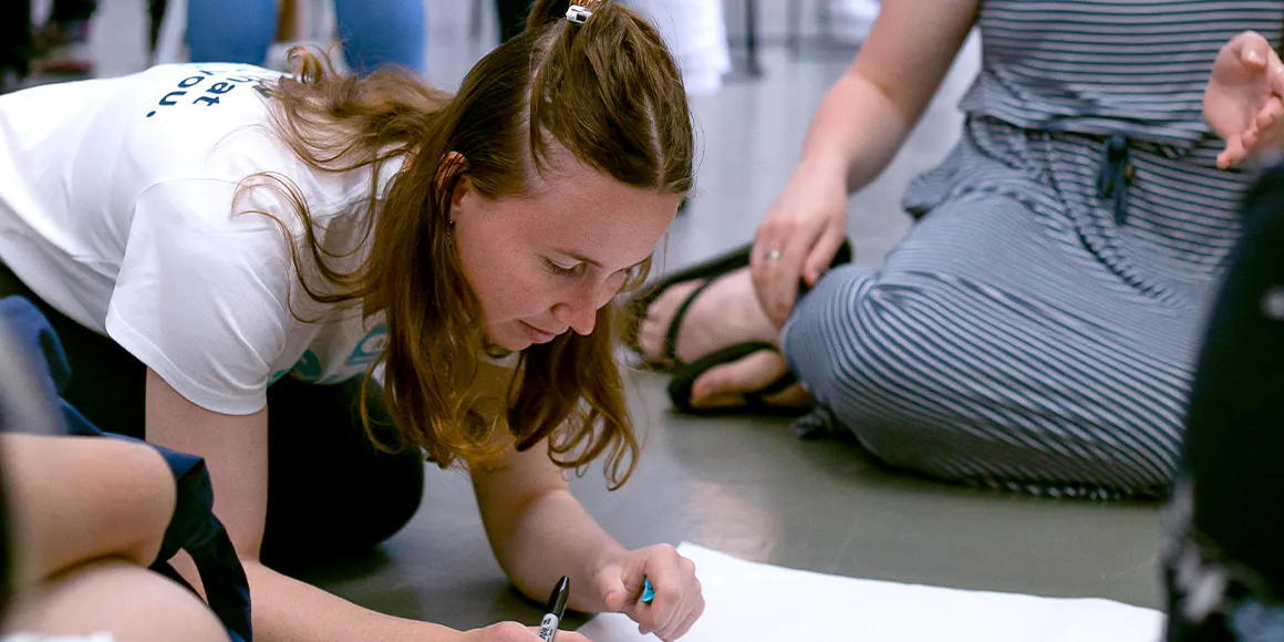A woman is writing on a large piece of paper. She's kneeling on the floor.