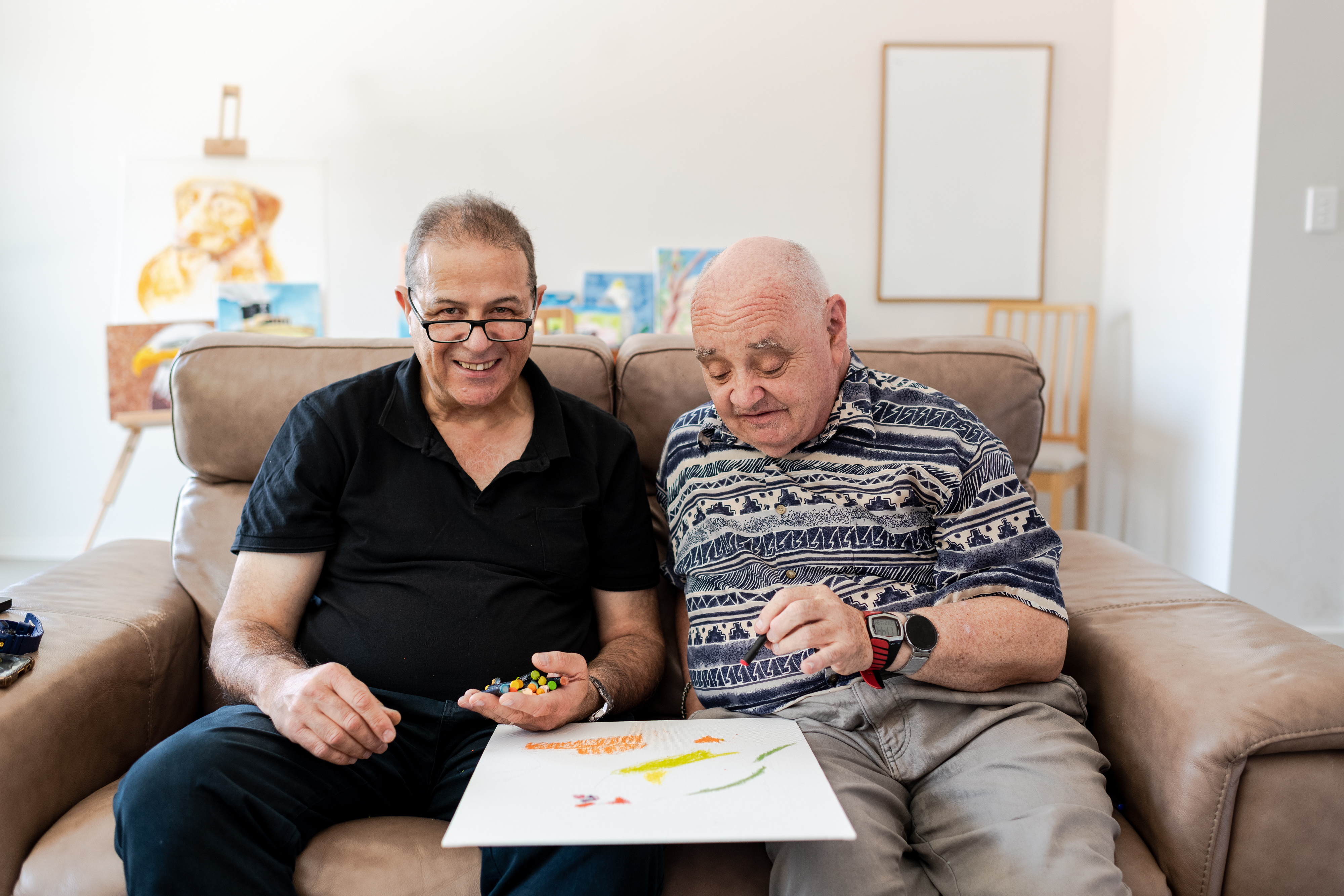 Daniel and Graeme are sitting on a brown couch doing art together with oil pastels. Daniel has short brown hair, wears reading glasses, a black collared shirt and navy trousers. Graeme has short white hair and is wearing two digital watches, a blue and white patterned shirt and grey trousers. 