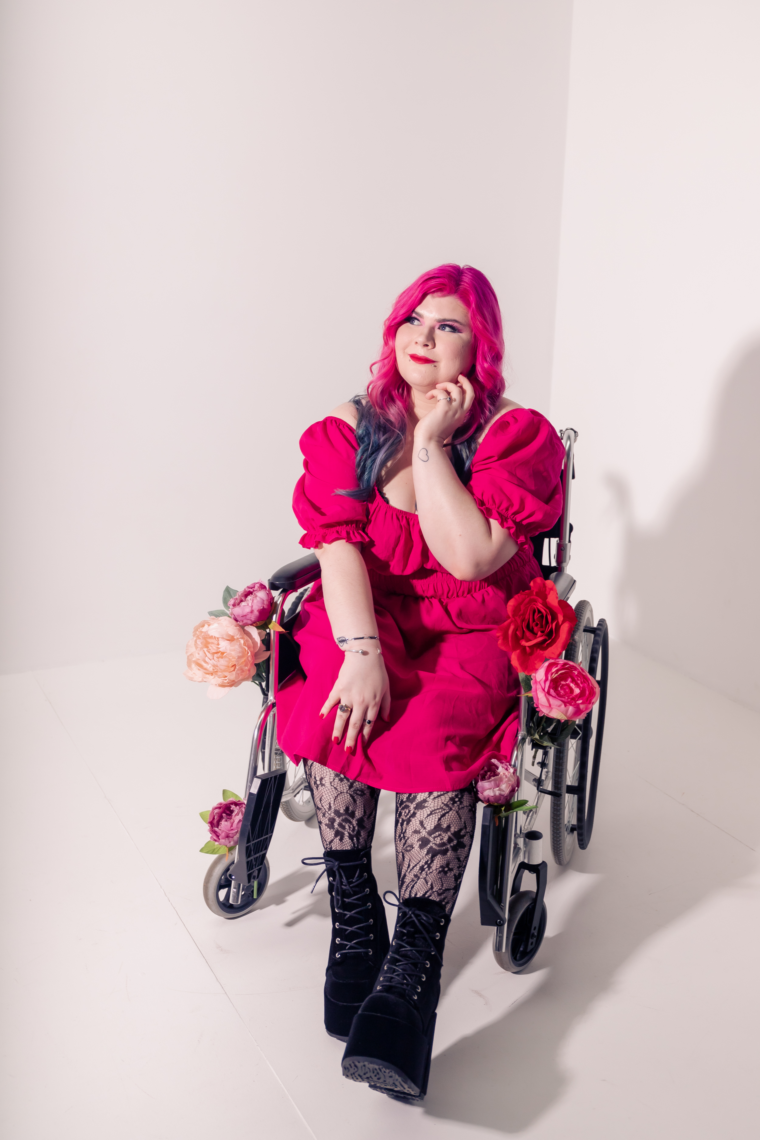 Zoe Simmons, a disability advocate and writer, is wearing a bright pink dress, floral fishnet stockings, and black heeled ankle boots. She has pale skin, long pink and blue hair, and bright makeup. She’s sitting in a silver and black manual wheelchair, adorned in fake flowers.