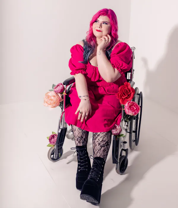 Zoe Simmons, a disability advocate and writer, is wearing a bright pink dress, floral fishnet stockings, and black heeled ankle boots. She has pale skin, long pink and blue hair, and bright makeup. She’s sitting in a silver and black manual wheelchair, adorned in fake flowers.
