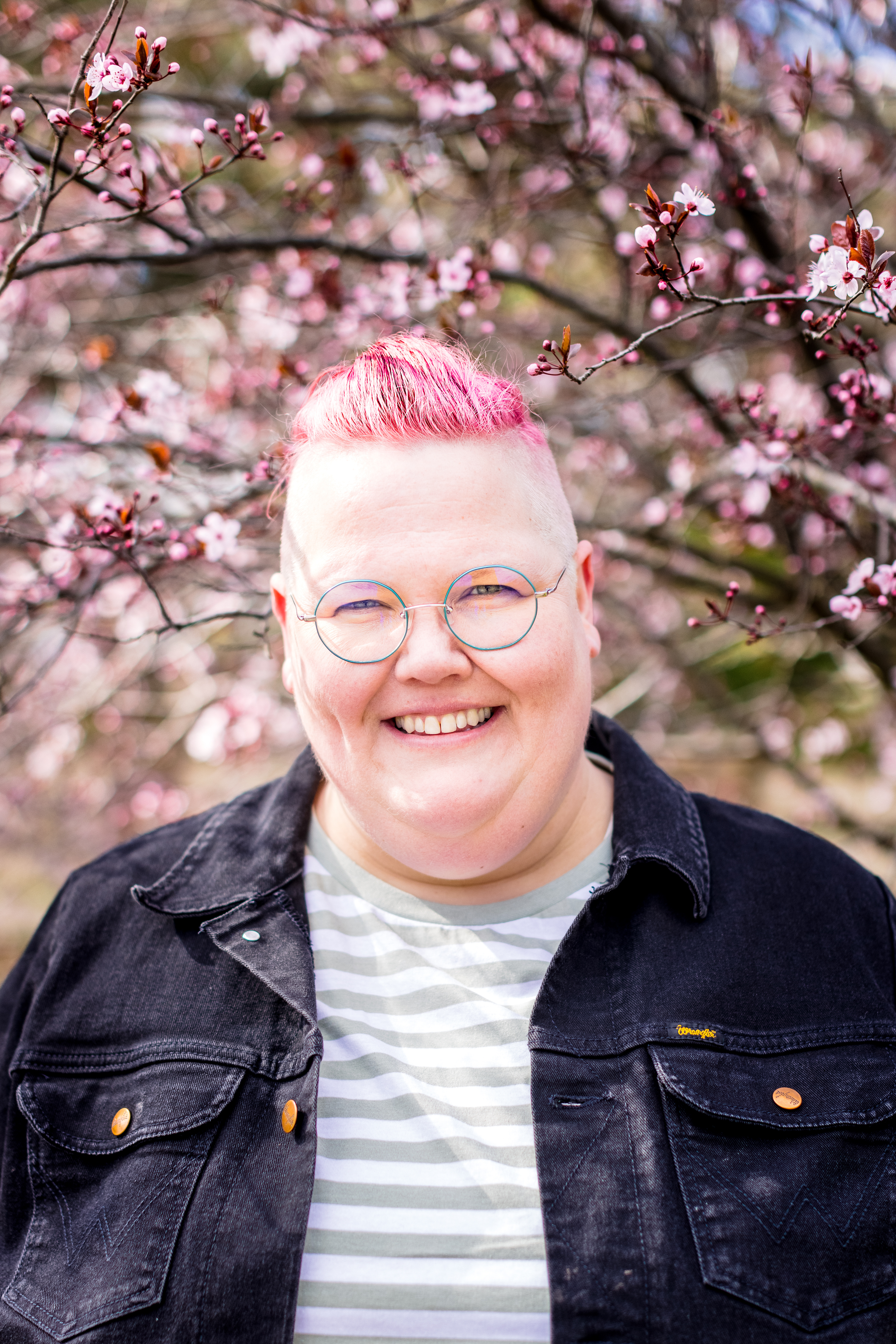 Jasper has bright pink hair and wears glasses. They're standing under a tree that has pink blossoms. 