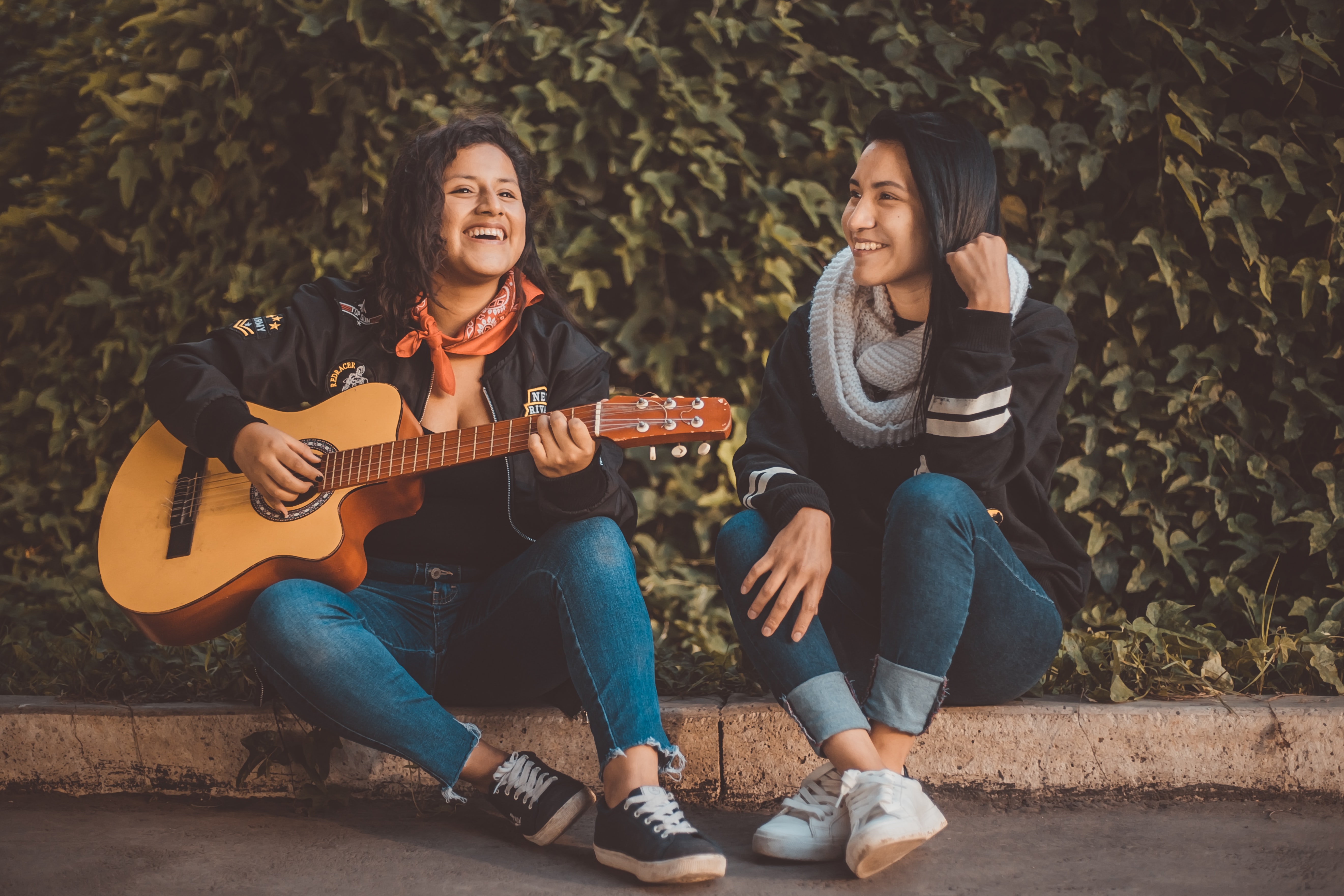 Two women sit side by side on the ground outside. One is playing a guitar. They're both smiling. 