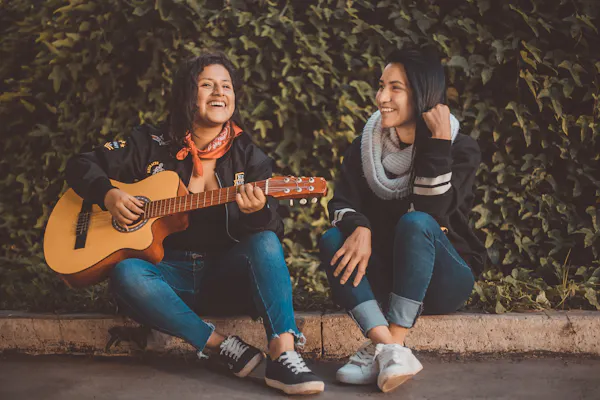 Two women sit side by side on the ground outside. One is playing a guitar. They're both smiling. 
