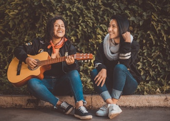 Two women sit side by side on the ground outside. One is playing a guitar. They're both smiling.
