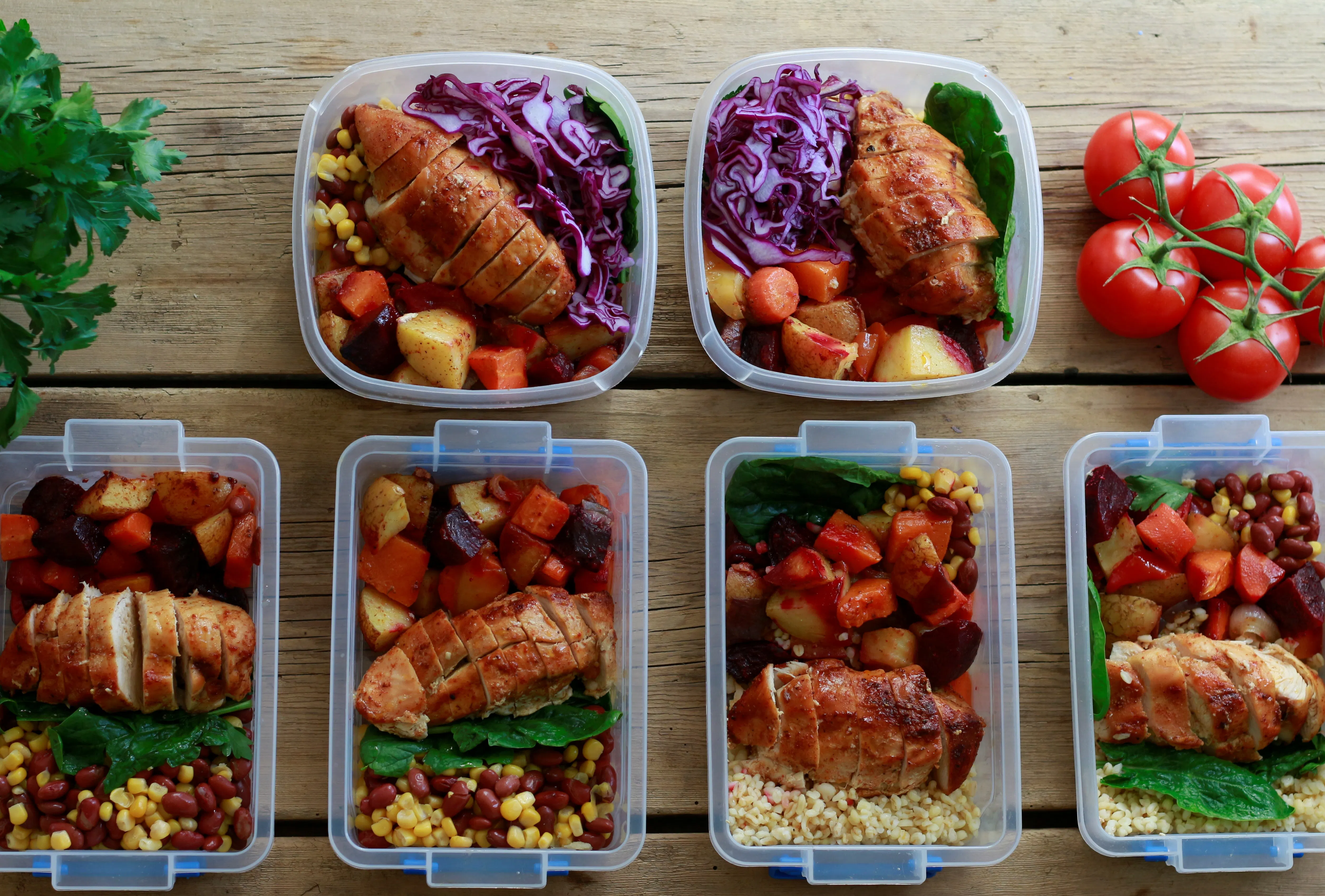 Colourful meals are laid out in clear plastic containers. They contain chicken, purple cabbage, potatoes, carrots, corn, and beans.