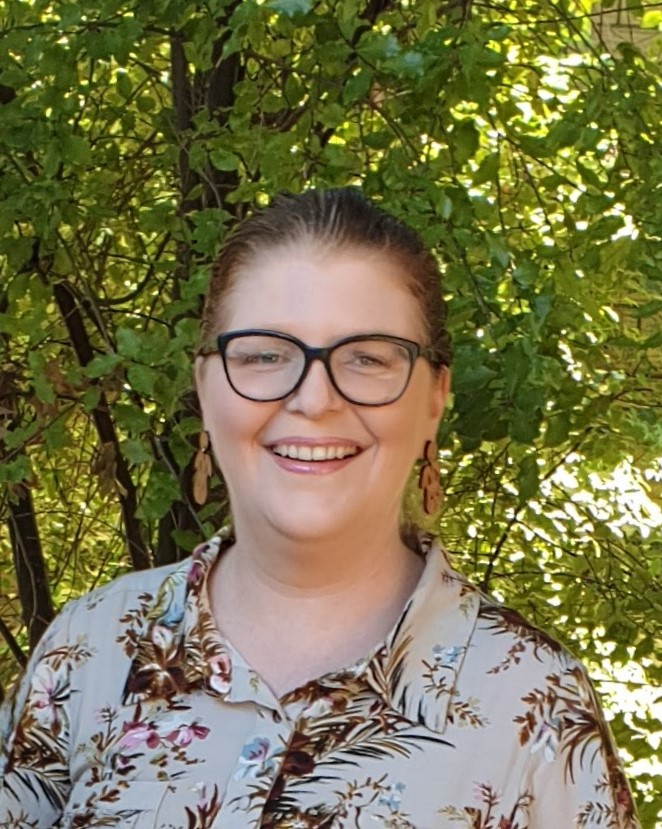 Libby Lake wears glasses and wooden earrings. She smiles and is standing in front of a leafy tree. 