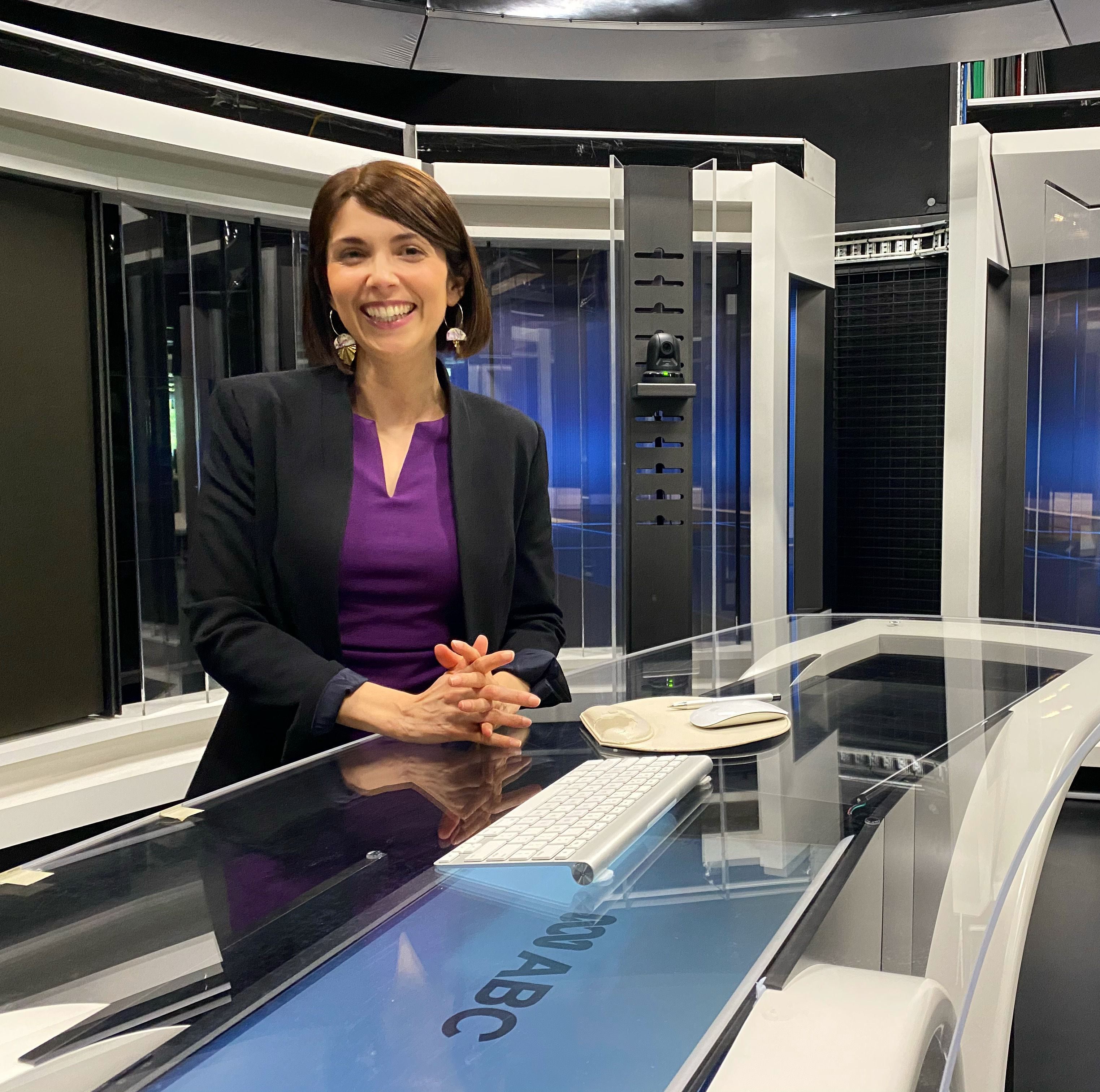 Natalia Hodgins stands behind a news desk and smiles. She's wearing a purple shirt and black blazer. 