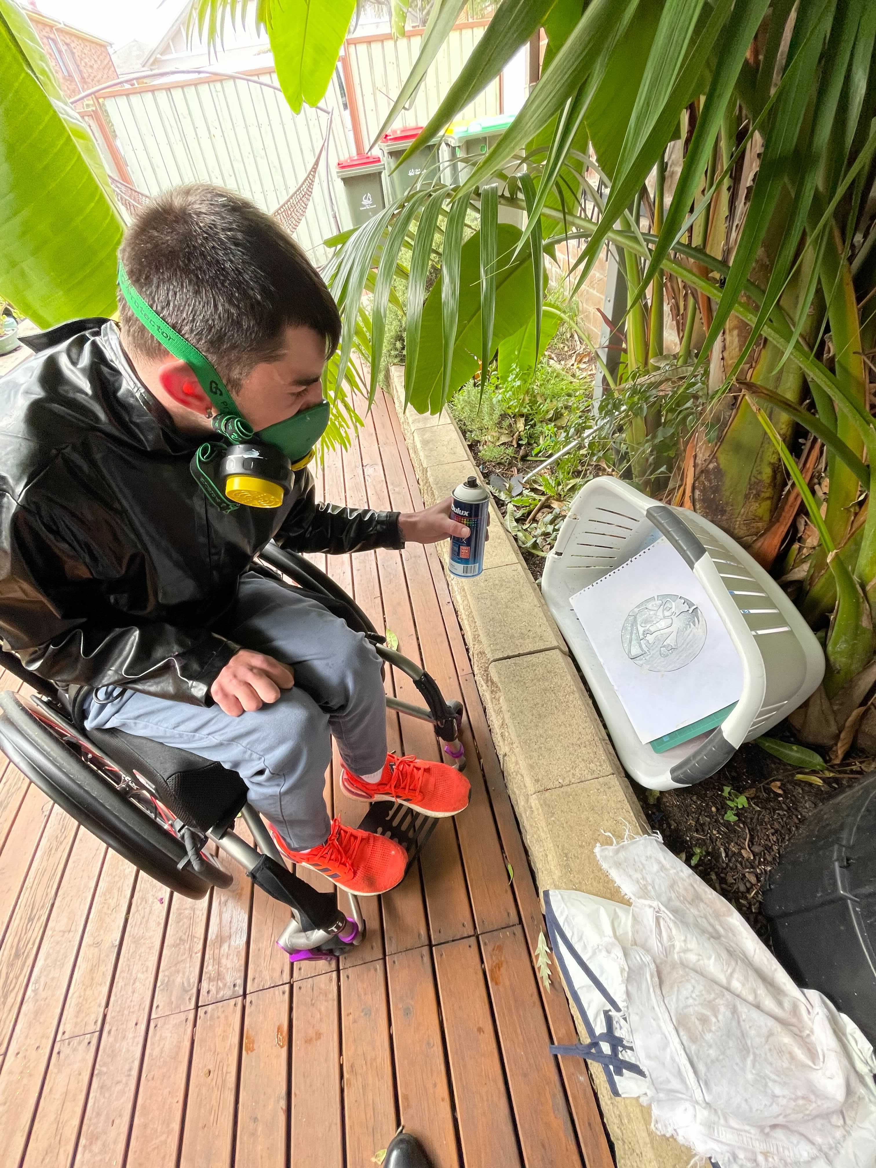 Andy is outside, spray painting a design onto paper. The paper is in a washing basket and Andy is wearing a gas mask.