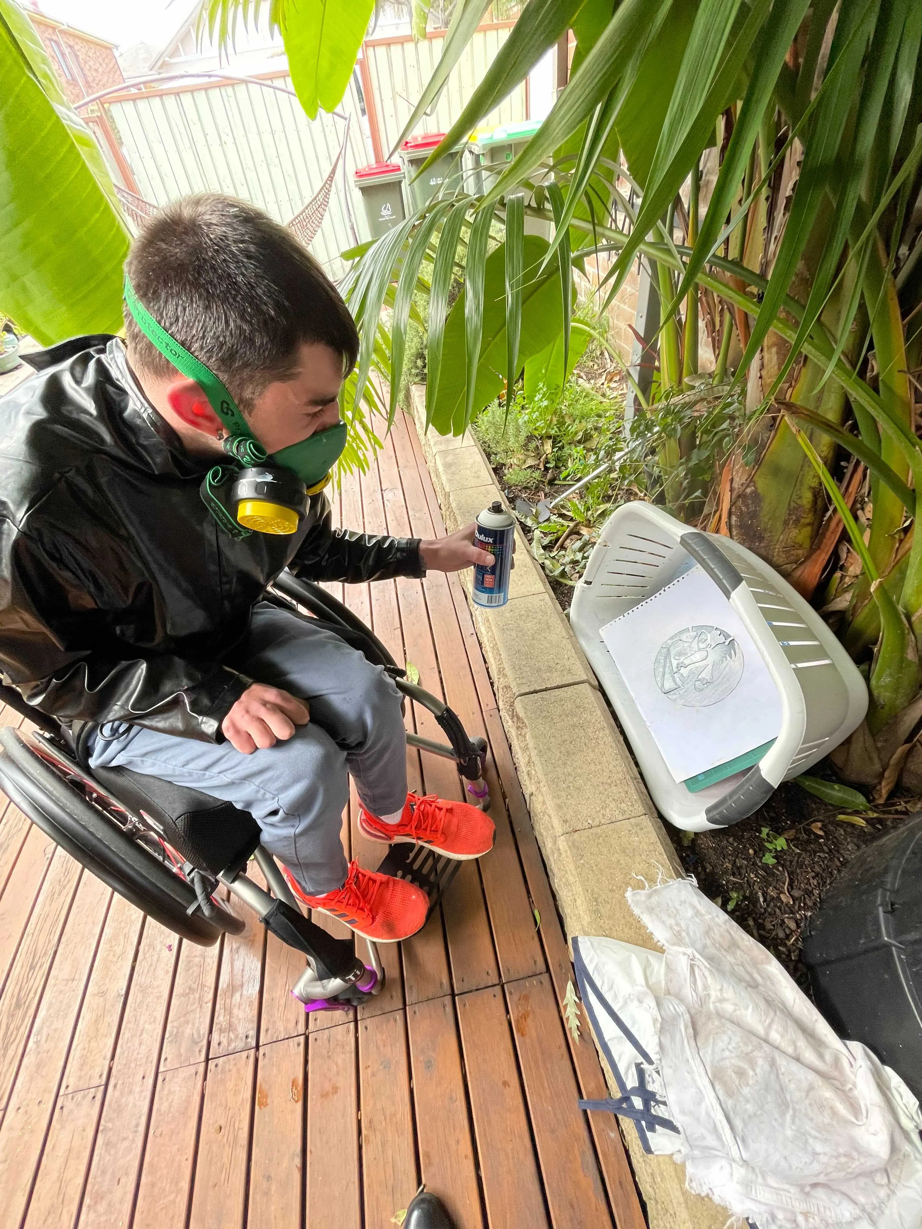 Andy is outside, spray painting a design onto paper. The paper is in a washing basket and Andy is wearing a gas mask.
