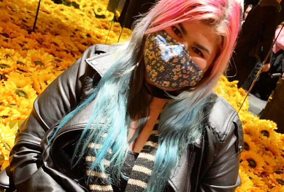 Zoe has long pink and blue hair, and is seated. She's wearing a PPE mask. A massive bunch of sunflowers surround the chair.