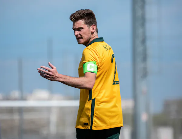 Australian Paralympian, Benny Roche, waves his left hand. He's wearing a green and gold soccer uniform and has an arm band with the letter 'c' on it.