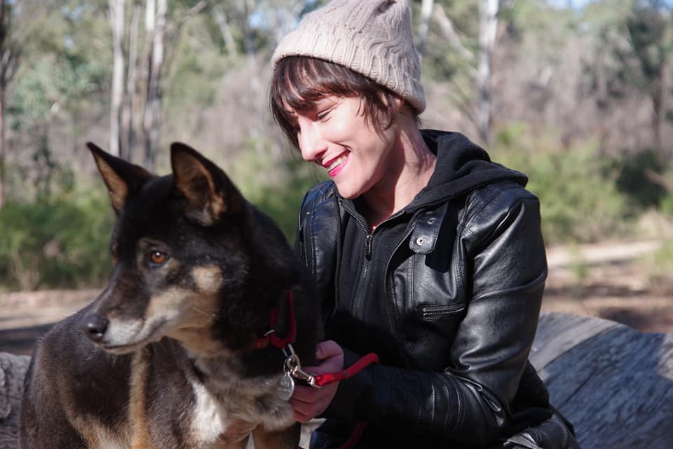 Elena wears a beanie and leather jacket. She's petting a dog and is smiling. Elena sits on a tree log.