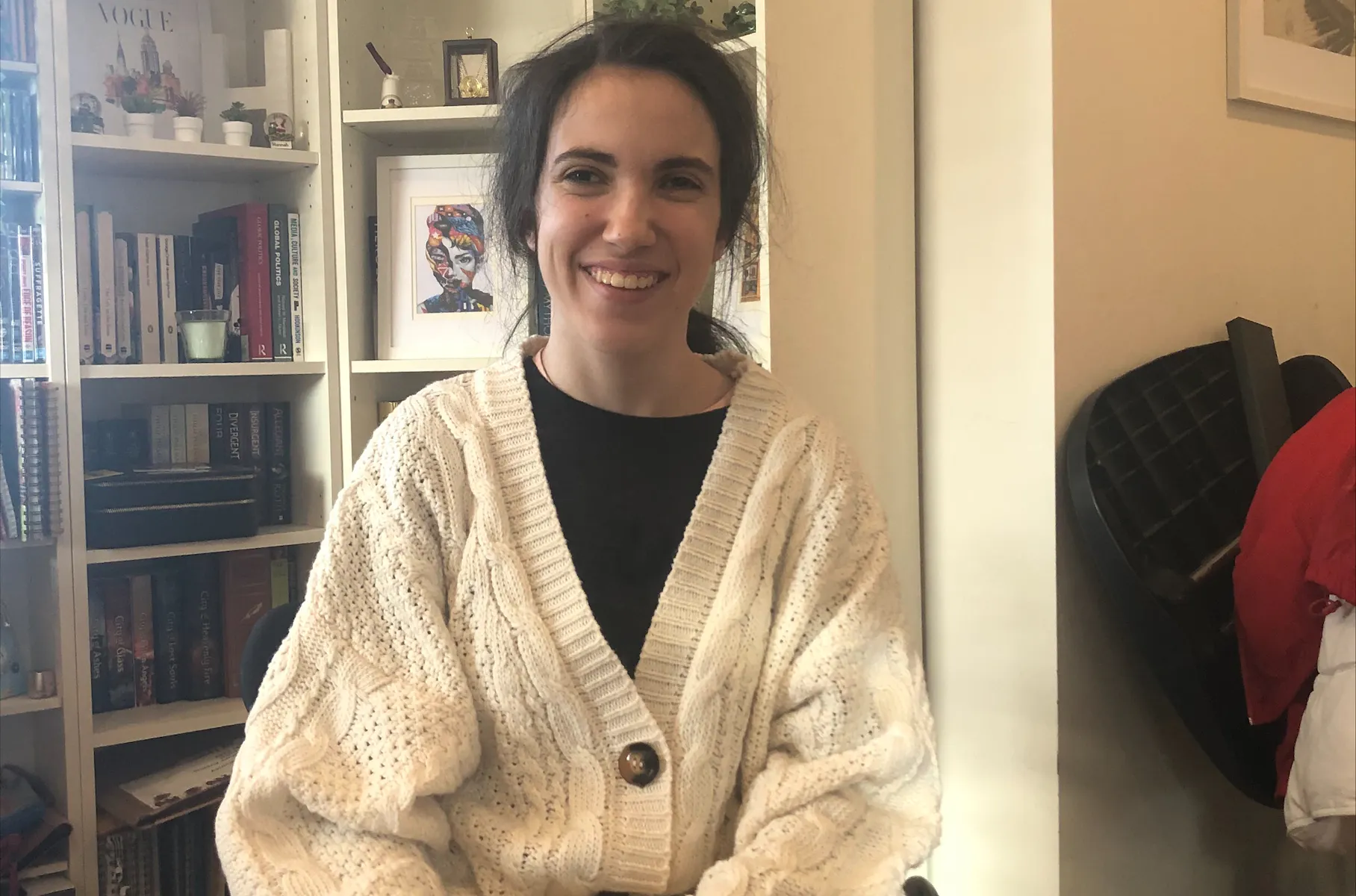 Hannah has her hands resting on her knees and is smiling. She's wearing a large cardigan. Hannah is a wheelchair user.