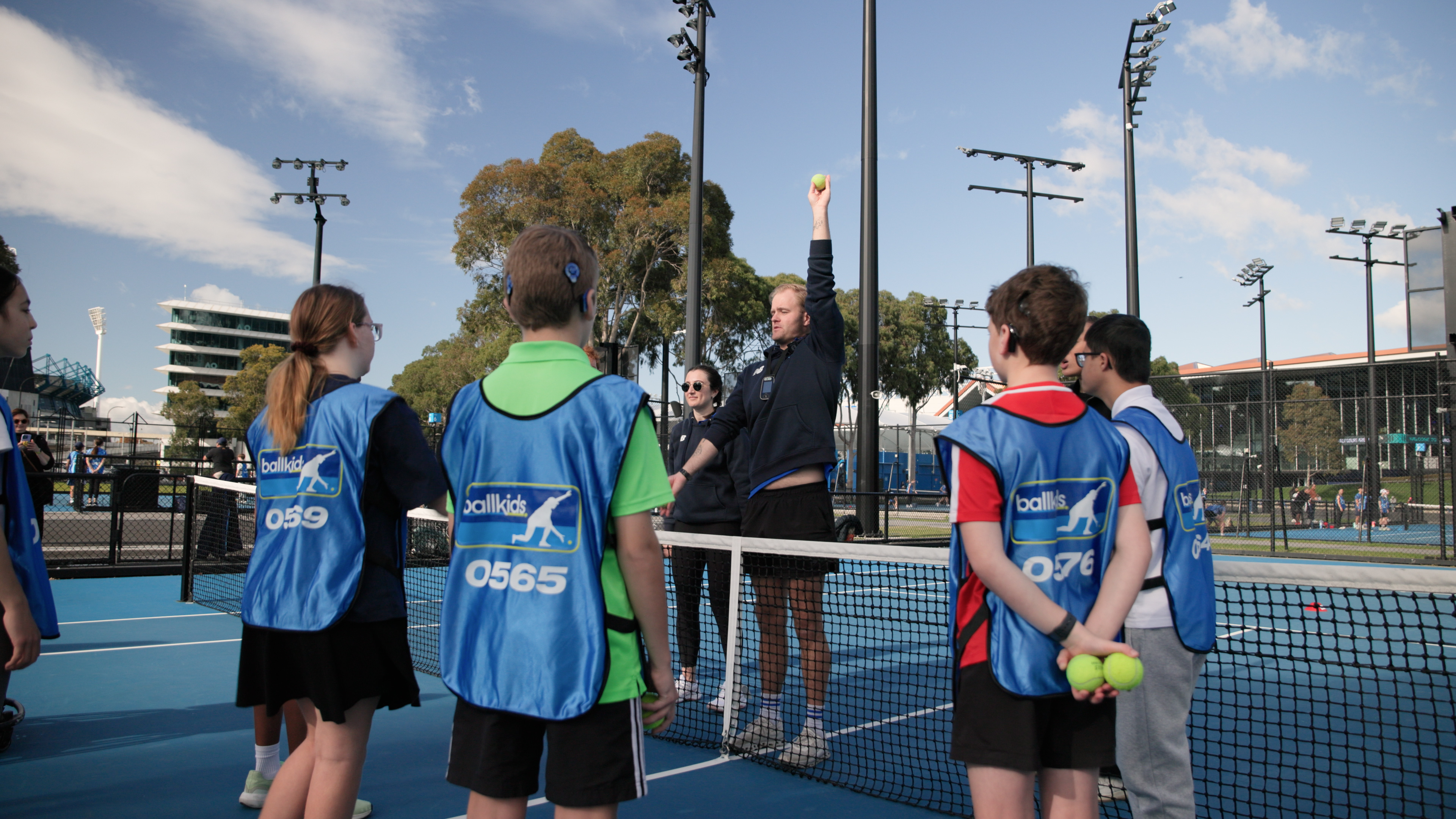 Daniel, a member of Tennis Australia’s Ballkid Operations team, is holding a tennis ball high with his arm fully extended. He's demonstrating this action to a group of ballkids gathered around him on the tennis court, all dressed in their blue smocks with the word 'Ballkid' printed on it.