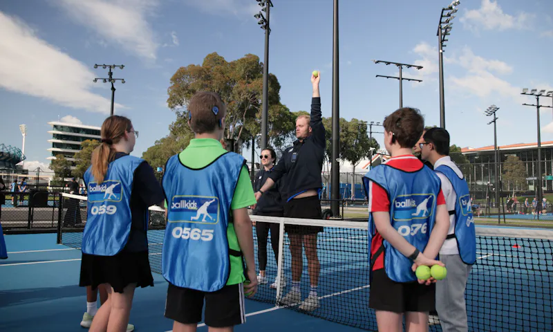 Daniel, a member of Tennis Australia’s Ballkid Operations team, is holding a tennis ball high with his arm fully extended. He's demonstrating this action to a group of ballkids gathered around him on the tennis court, all dressed in their blue smocks with the word 'Ballkid' printed on it.