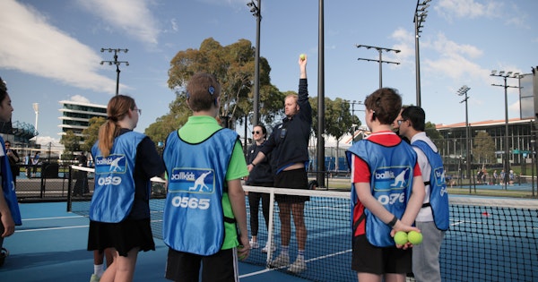 Daniel, a member of Tennis Australia’s Ballkid Operations team, is holding a tennis ball high with his arm fully extended. He's demonstrating this action to a group of ballkids gathered around him on the tennis court, all dressed in their blue smocks with the word 'Ballkid' printed on it.