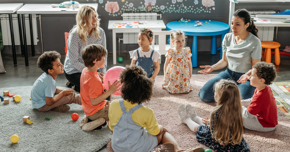 A group of young children sit in a circle with two teachers. They're at a supported playgroup and have toys surrounding them. 