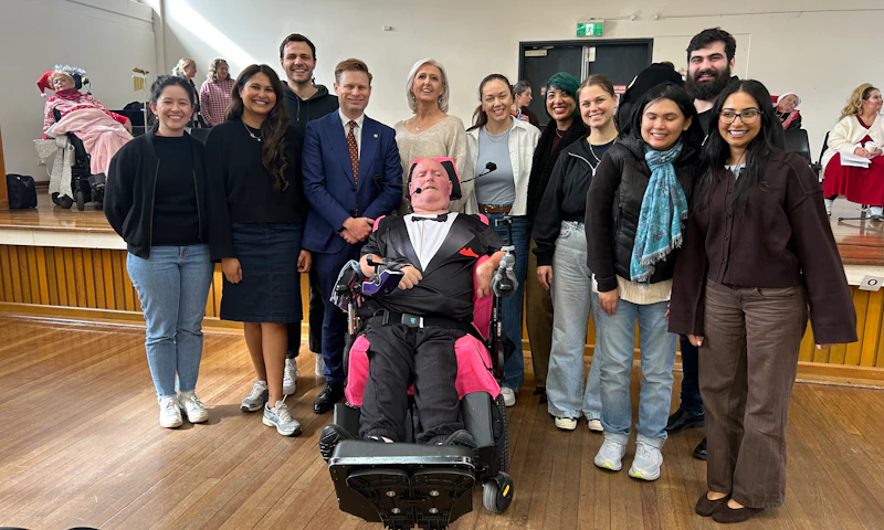 Marc, a Hireup Impact Grant recipient, is wearing a tuxedo and wireless headset microphone. He's surrounded by Hireup staff members who are dressed in winter clothing, smiling. They're in a community hall for a choir performance that Marc is conducting.