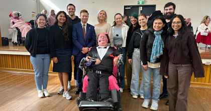 Marc, a Hireup Impact Grant recipient, is wearing a tuxedo and wireless headset microphone. He's surrounded by Hireup staff members who are dressed in winter clothing, smiling. They're in a community hall for a choir performance that Marc is conducting.