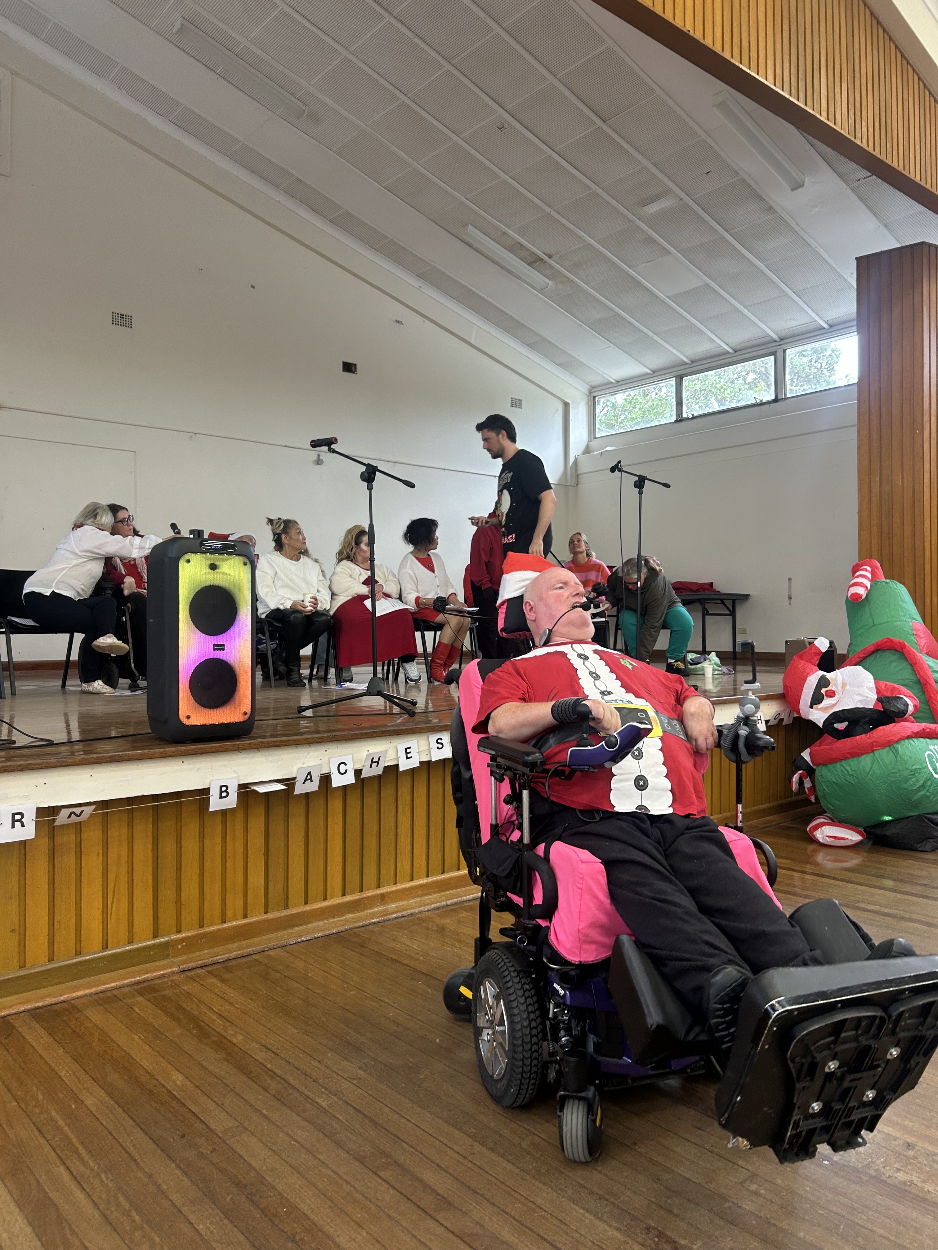 Marc, a Hireup Impact Grant recipient, is centre stage at a community hall. Marc, a wheelchair user, is speaking into a microphone and is wearing a Santa Claus suit. He's conducting a community choir performance that has a Christmas in July theme. 