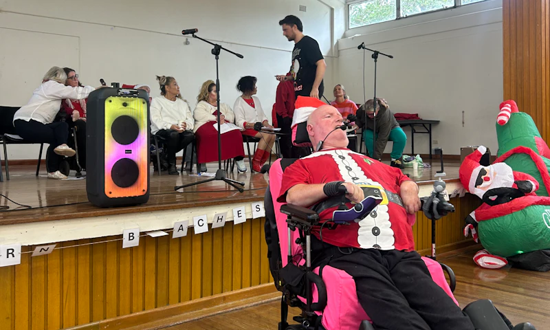 Marc, a Hireup Impact Grant recipient, is centre stage at a community hall. Marc, a wheelchair user, is speaking into a microphone and is wearing a Santa Claus suit. He's conducting a community choir performance that has a Christmas in July theme.