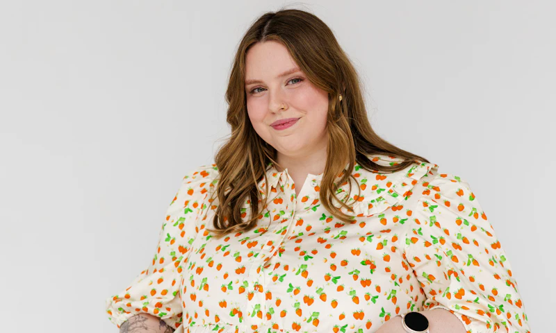 Jasmin Scriven, a disability advocate and content creator, has shoulder-length brown hair and wears a long strawberry-print dress. Jasmin is a wheelchair user.