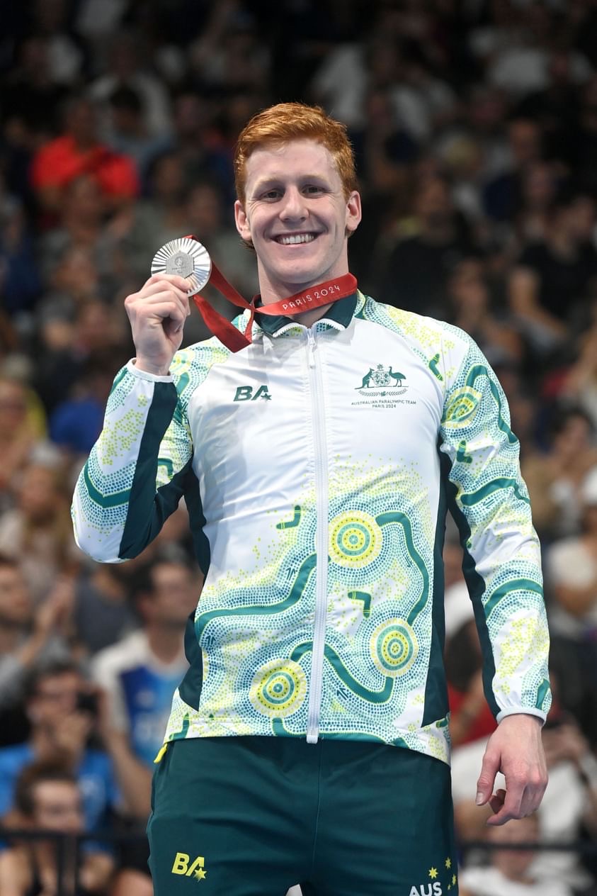 Australian Paralympian Col Pearse is holding up a silver medal, grinning. He's in an arena with thousands of spectators. Col is wearing a white jacket that has a green and gold Indigenous artwork printed onto it, plus dark green tracksuit pants. 