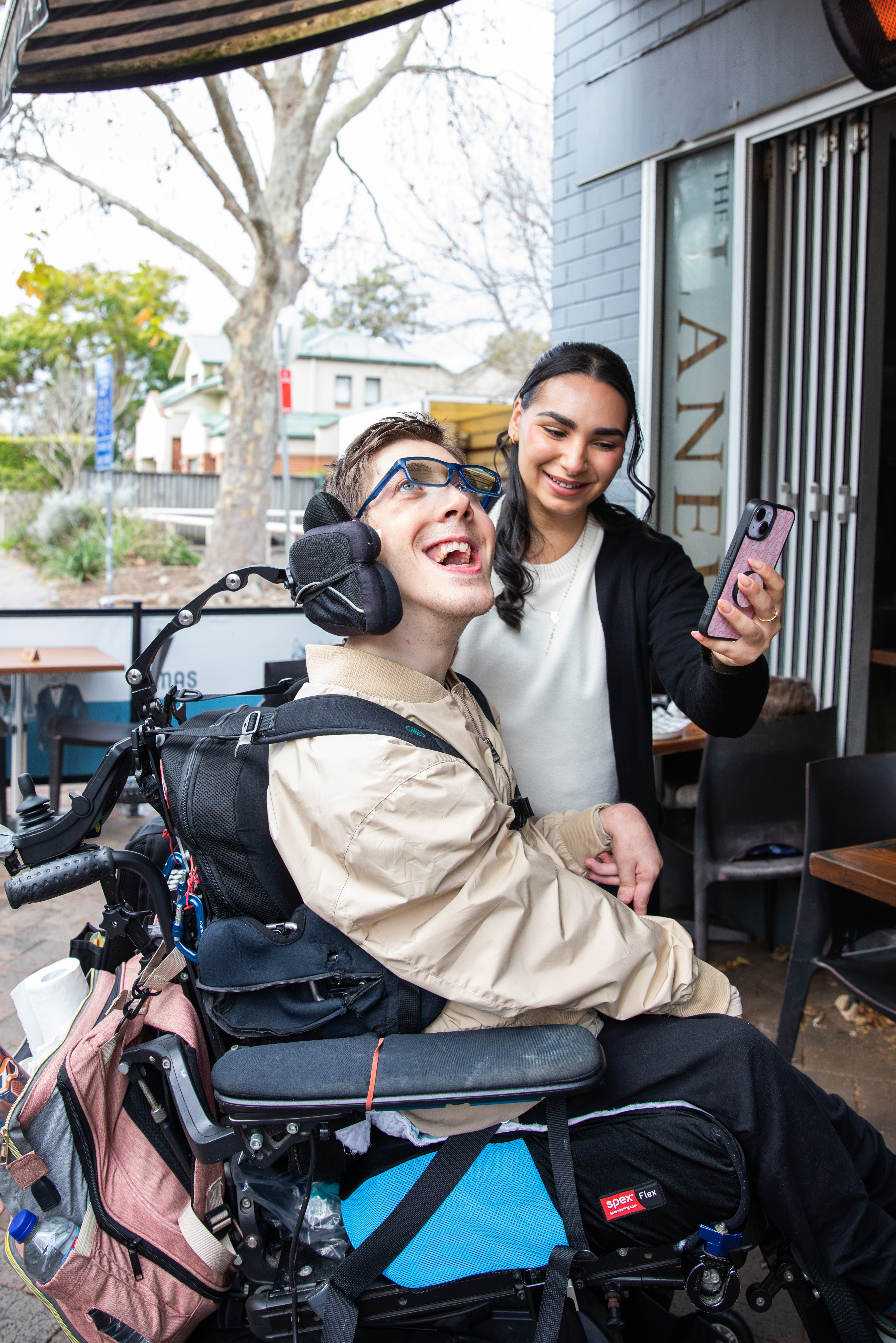 Hireup client Al laughing with his support worker, Bec. Al, who is a wheelchair user, has short brown hair and wears a beige jacket and glasses. He's looking at a phone that Bec is holding in front of him. Bec has long dark hair and wears a top and cardigan.