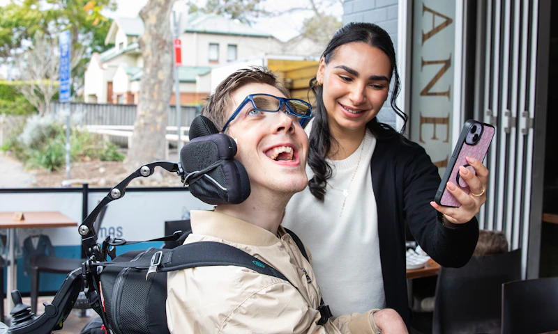 Hireup client Al laughing with his support worker, Bec. Al, who is a wheelchair user, has short brown hair and wears a beige jacket and glasses. He's looking at a phone that Bec is holding in front of him. Bec has long dark hair and wears a top and cardigan.
