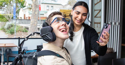 Hireup client Al laughing with his support worker, Bec. Al, who is a wheelchair user, has short brown hair and wears a beige jacket and glasses. He's looking at a phone that Bec is holding in front of him. Bec has long dark hair and wears a top and cardigan.