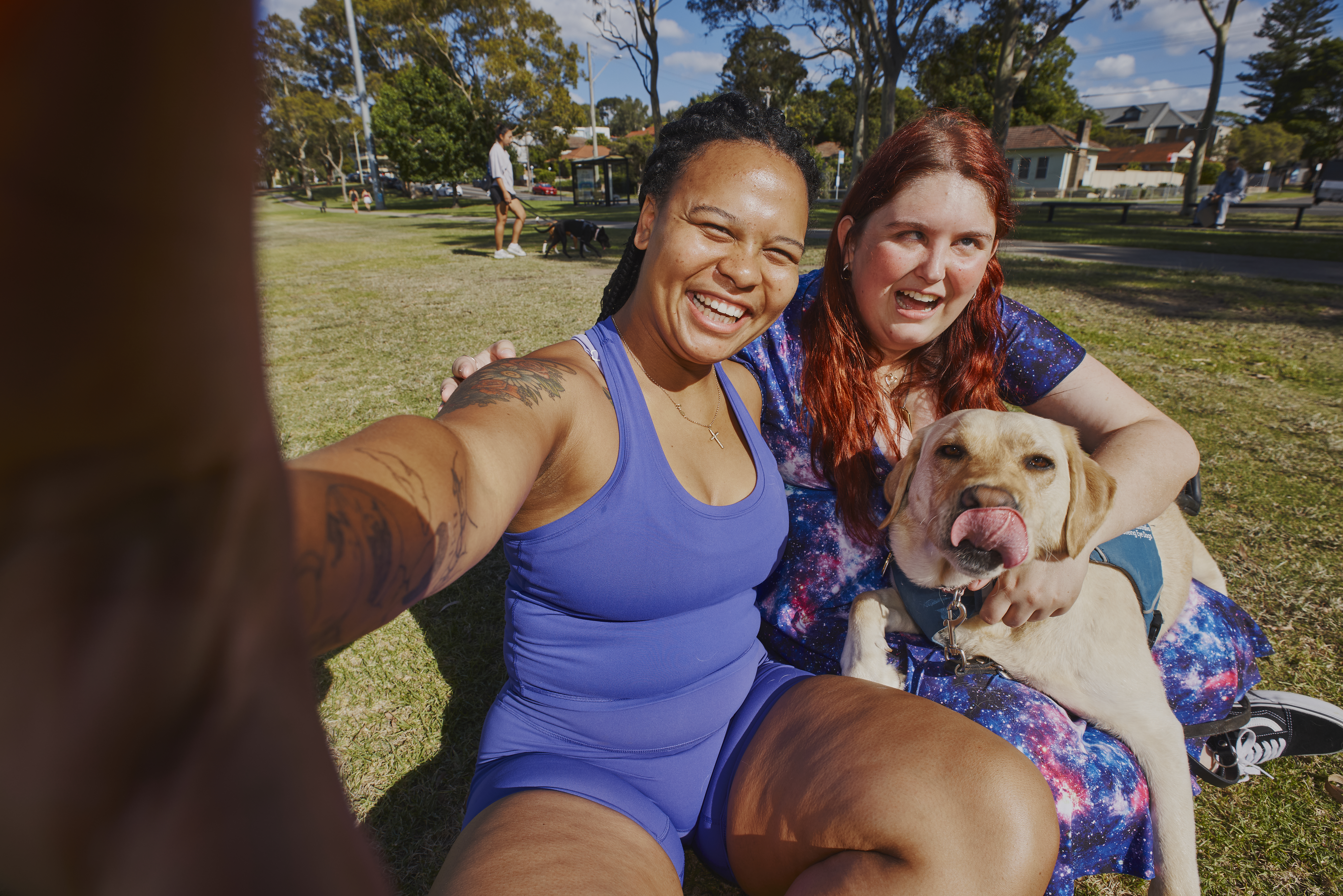 Kirsten and her Hireup support worker Paulla are in a park, smiling. Guide dog, Kimba, lays across Kirsten's lap. 