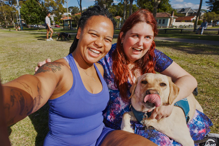 Kirsten and her Hireup support worker Paulla are in a park, smiling. Guide dog, Kimba, lays across Kirsten's lap.