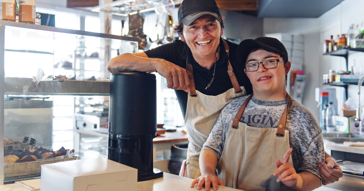 Two men are standing at a cafe counter dressed in aprons and hats, smiling.