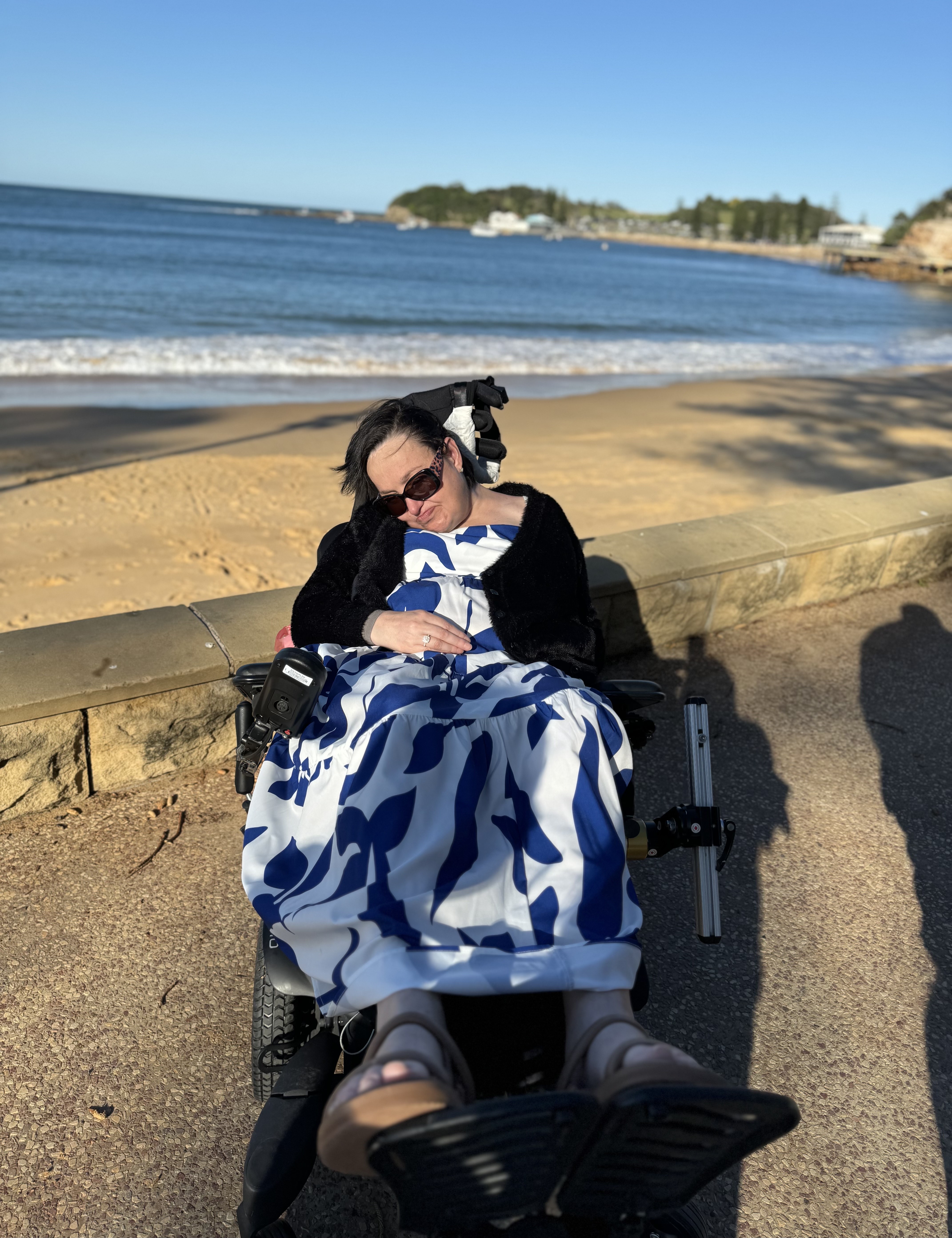 Judy is soaking up the sunshine at the beach. She is wearing sunglasses, a blue and white maxi dress, and cardigan. Judy uses a wheelchair. 