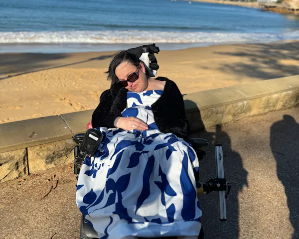 Judy is soaking up the sunshine at the beach. She is wearing sunglasses, a blue and white maxi dress, and cardigan. Judy uses a wheelchair.