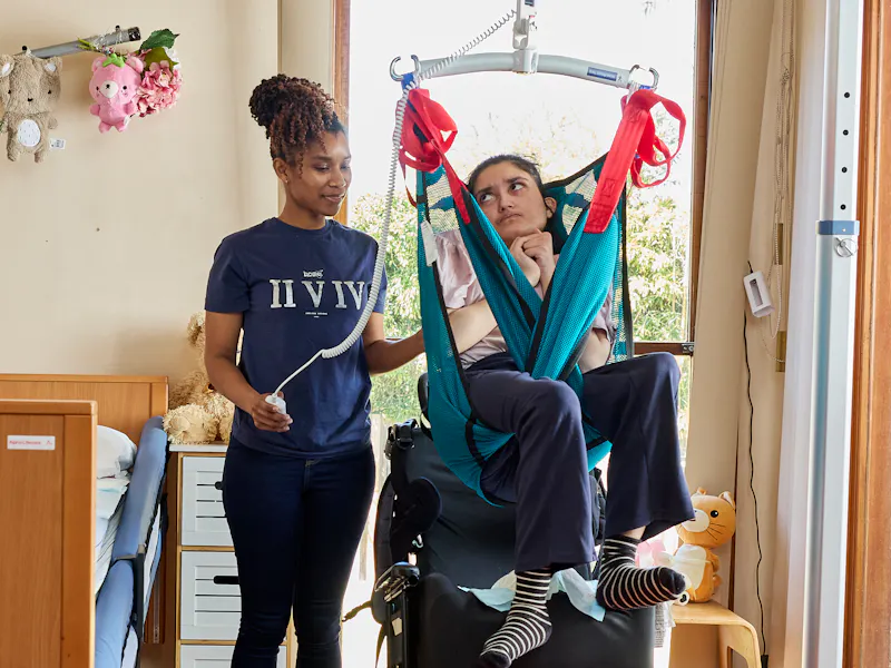 Rifa and her support worker Mariam are in a bedroom. Rifa is wearing a pink top and is looking at Mariam. Mariam is gently touching Rifa's arm and is smiling.