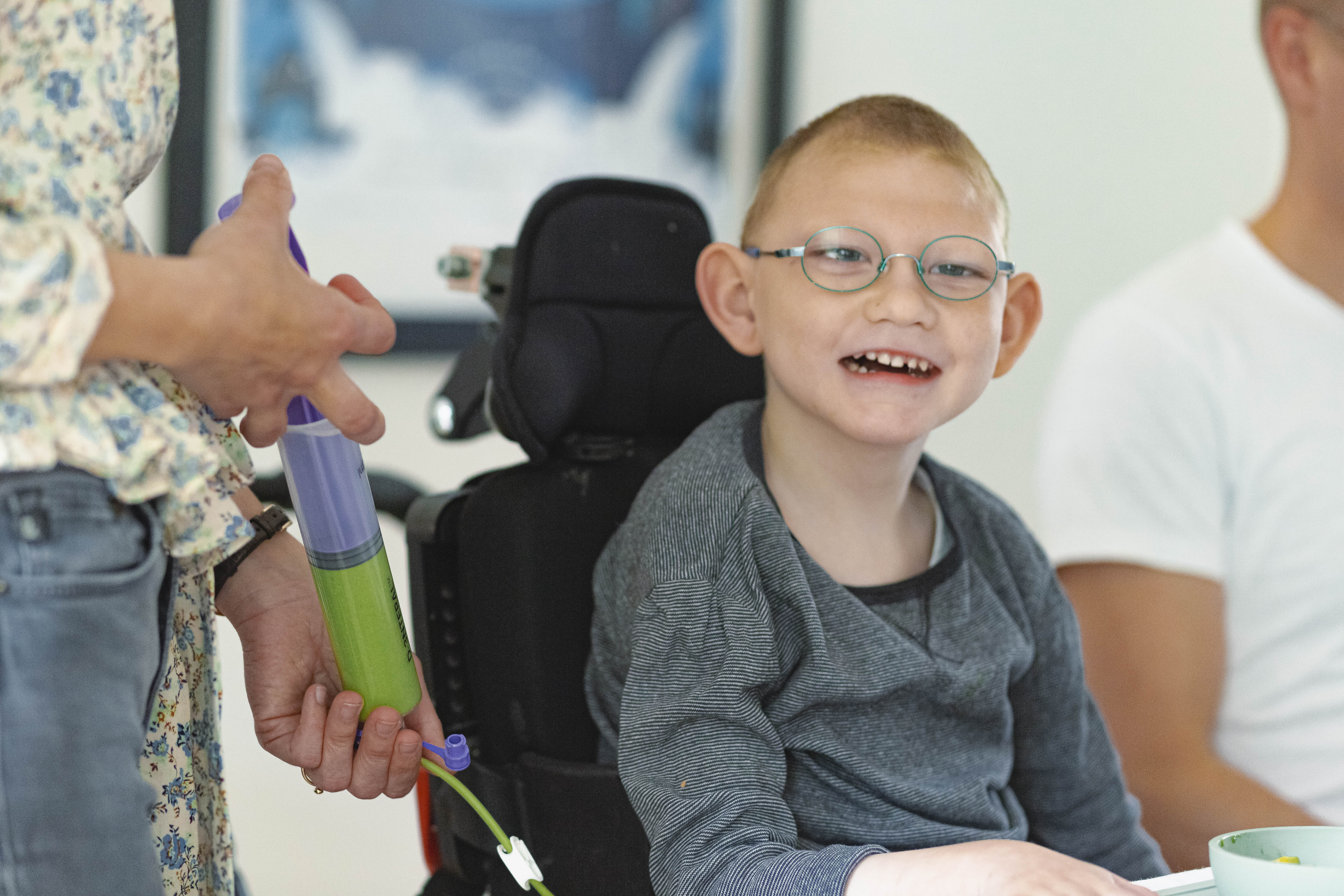 A young boy sits at the kitchen table in his wheelchair. He's smiling as he gets tube fed. He wears glasses and a grey jumper. 