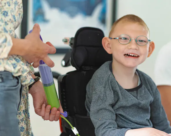 A young boy sits at the kitchen table in his wheelchair. He's smiling as he gets tube fed. He wears glasses and a grey jumper.