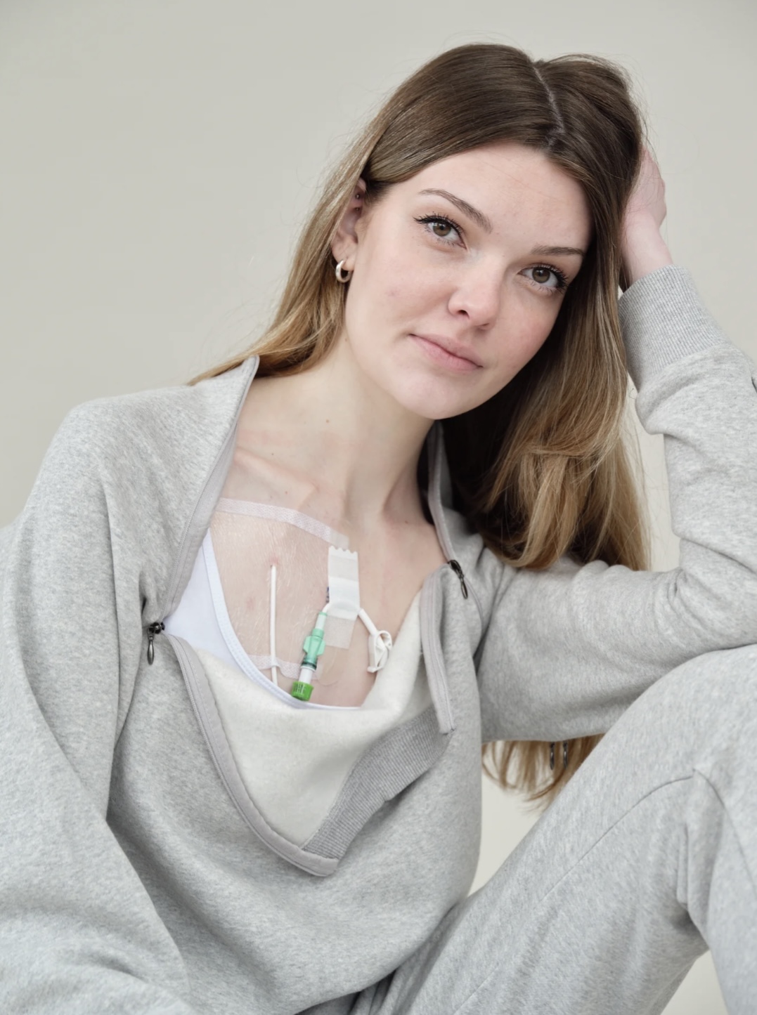 Celia gently smiles as she rests her head on her arm. She is wearing a grey jumpsuit, and her top is unzipped, revealing feeding tubes and tape across her chest.