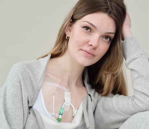Celia gently smiles as she rests her head on her arm. She is wearing a grey jumpsuit, and her top is unzipped, revealing feeding tubes and tape across her chest.