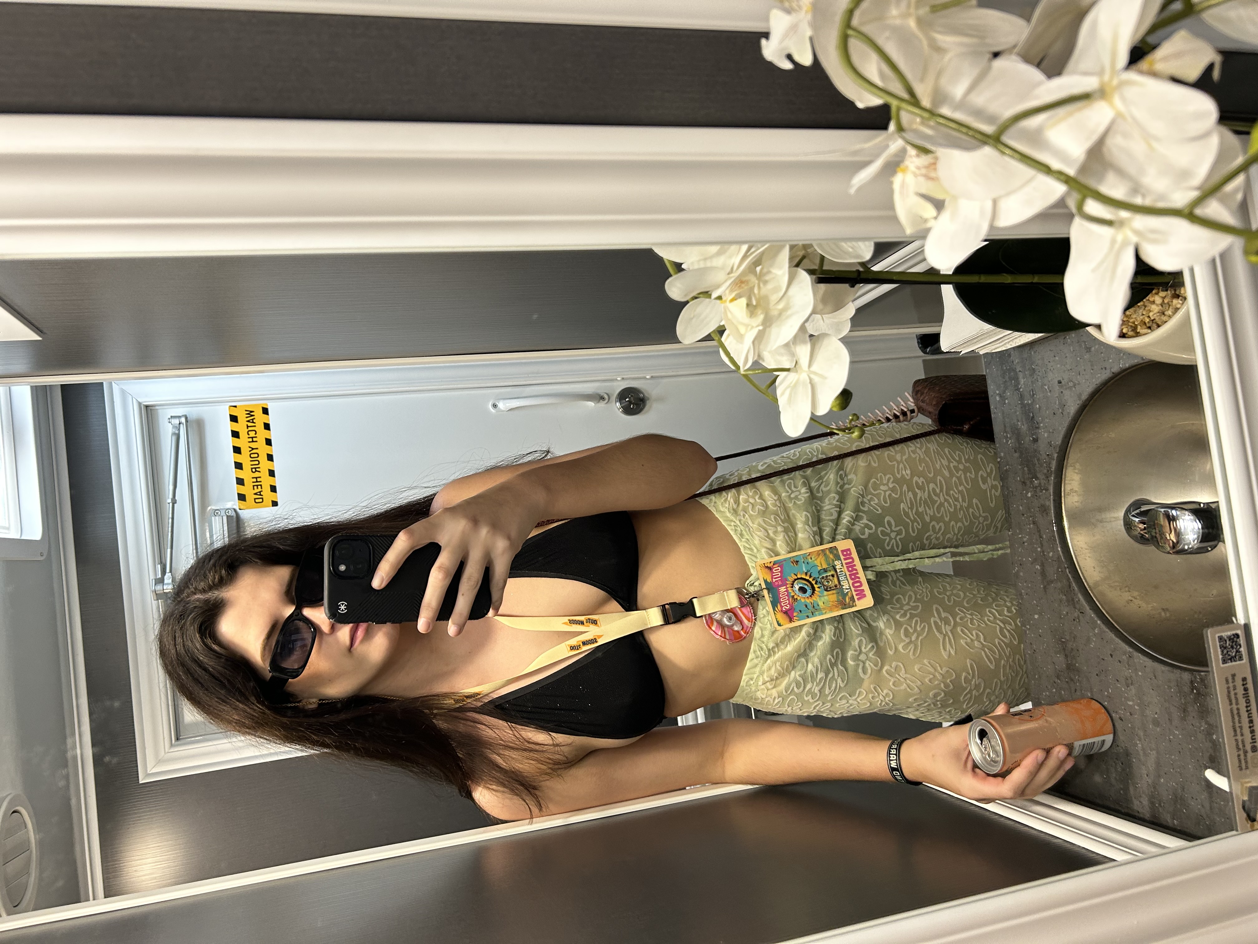 Elli is taking a selfie in a bathroom mirror. She's wearing a bikini top, a sheer skirt and sunglasses. Elli has a feeding-tube pad.