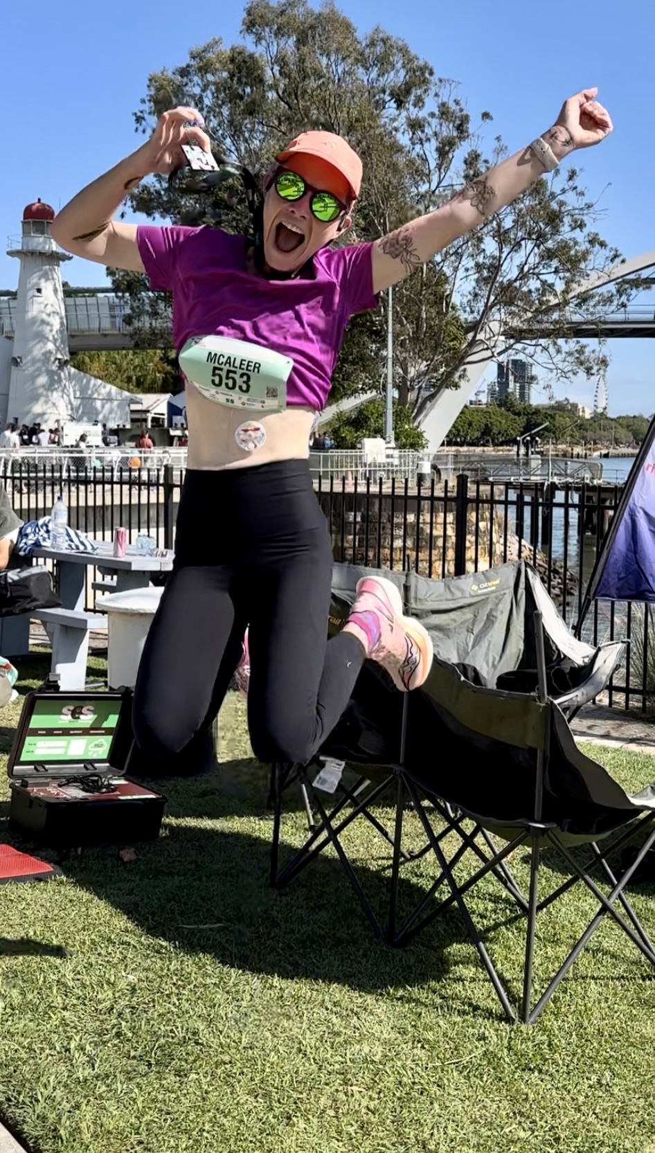 Juliette is leaping into the air, proudly wearing her marathon bib and holding up her medal in celebration. Juliette wears a tube-feeding pad.