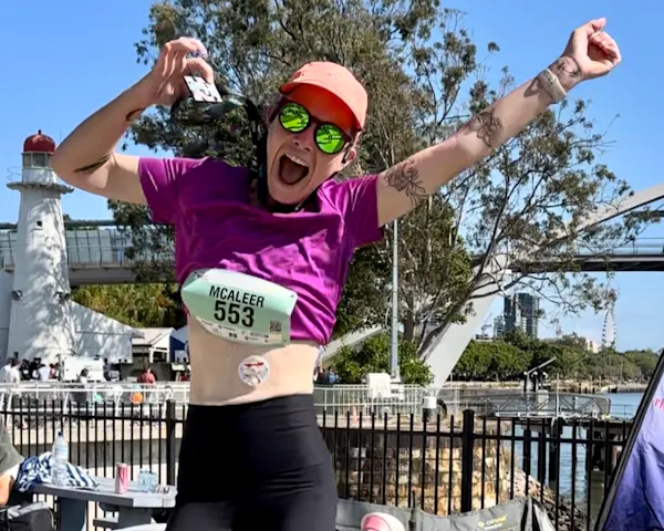 Juliette is leaping into the air, proudly wearing her marathon bib and holding up her medal in celebration. Juliette wears a tube-feeding pad.